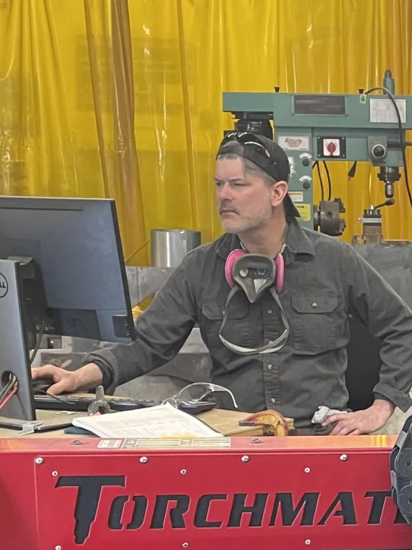 a white man with a ball cap on backwards wearing a respirator around his neck sitting at a computer