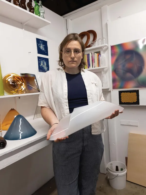 a person with shoulder length light brown hair wearing glasses holding a rolled white rolled piece of glass in front of them