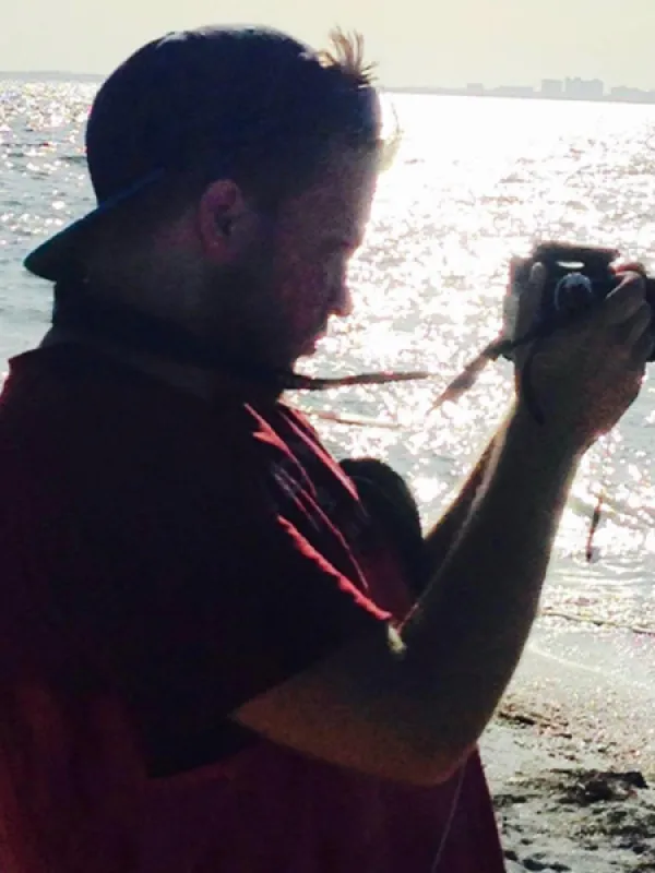 a person wearing  reversed baseball cap and a red shirt stands on shoreline capturing photos, 